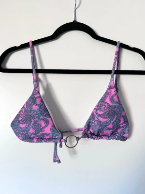 Mönstrad bikiniöverdel med ringdetalj - Snygg bikiniöverdel i rosa, grå och turkos med ett unikt mönster. Den har en dekorativ ringdetalj framtill och justerbara axelband för perfekt passform. Perfekt för sommarens stranddagar! Storlek L men liten i kuporna och passar bra till S/M. 