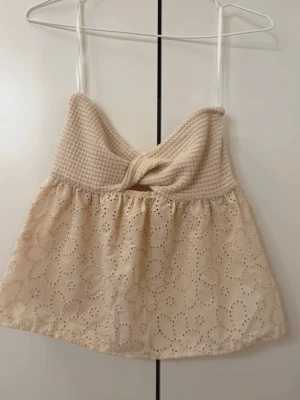Beige spetsig linnetopp med twist - Söt beige linnetopp med twistad detalj framtill. Överdelen är stickad och nederdelen har broderade blommor i spets. Perfekt för sommarens varma dagar.
