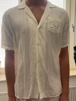 Vit linneskjorta med kort ärm - Säljer en luftig vit skjorta i linne med korta ärmar och klassisk krage. Skjortan har en bröstficka och knäppning framtill. Perfekt för varma dagar och enkel att styla till olika outfits. Aldrig använd eftersom den tyvärr har blivit för liten! Nypris-1000kr/ mitt pris- 250kr!