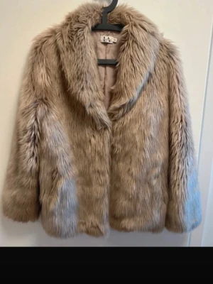 Beige fluffig fuskpälsjacka LA - Säljer en beige fuskpälsjacka med bred krage och långa ärmar. Jackan har en mjuk och fluffig känsla, stängs framtill och har en klassisk rak passform. Perfekt för dig som vill ha en trendig och varm look under kalla dagar. Linn Ahlborg kollektion
