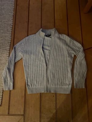 Ljusgrå ribbad kofta från Dressmann - Snygg ljusgrå ribbstickad kofta från Dressmann med dragkedja framtill och hög krage. Koftan har långa ärmar och breda muddar vid ärmslut och nederkant. Perfekt för lager på lager och enkel att matcha med olika outfits.