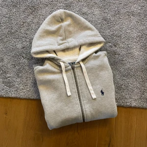 Grå hoodie från Polo Ralph Lauren - Säljer en stilren grå hoodie från Polo Ralph Lauren med dragkedja och två fickor framtill. Klassisk huva med vita snören och den ikoniska blå loggan broderad på bröstet. Perfekt för en avslappnad och trendig look.