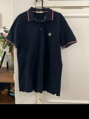 Marinblå pikétröja från Moncler - Snygg marinblå pikétröja från Moncler med klassisk krage och knappar framtill. Tröjan har korta ärmar och detaljer i rött, vitt och blått på krage och ärmslut samt Moncler-logga på bröstet. Perfekt för en stilren och sportig look.