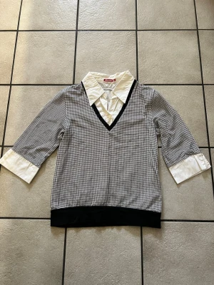 Rutig skjorta med v-ringning storlek M - Säljer en svartvit rutig skjorta från med v-ringad krage och vit klassisk skjortkrage. Skjortan har trekvartsärm med vita manschetter och svart ribbad nederkant. Perfekt för dig som gillar en preppy look.