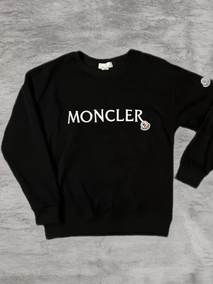Svart sweatshirt från Moncler - Stilren svart sweatshirt från Moncler med vit logotyptext på bröstet och patchdetalj på ärmen. Tröjan har rund halsringning, ribbade muddar och är tillverkad i mjukt bomullsmaterial. Perfekt för en avslappnad och trendig look.