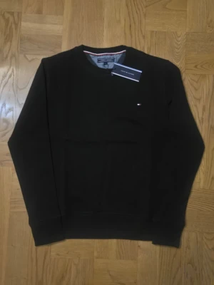 Svart sweatshirt från Tommy Hilfiger - Klassisk svart sweatshirt från Tommy Hilfiger med rund halsringning och diskret logga på bröstet. Tröjan har långa ärmar och ribbade muddar vid ärmslut och nederkant. Perfekt för en stilren och enkel look.