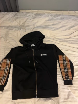 Svart Burberry hoodie med rutiga detaljer - Snygg svart hoodie från Burberry med dragkedja och huva. Ärmarna har klassiska Burberry-rutiga paneler i beige, rött och svart samt stor logga på ena ärmen. Tillverkad i mjukt material och har fickor framtill. Perfekt för dig som gillar streetwear och exklusiva märken.