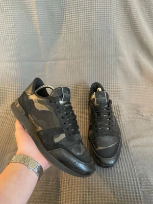 Valentino rockrunner  - Snygga svarta sneakers från Valentino med camo-mönster i olika nyanser av svart och grått. Skorna har en mix av mocka och läderdetaljer, samt svarta skosnören och en robust sula. Perfekta för dig som vill ha en stilren men ändå unik look.