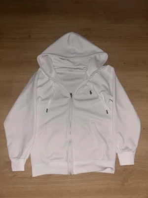 Vit hoodie från Polo Ralph Lauren - Snygg vit hoodie från Polo Ralph Lauren med dragkedja framtill och klassisk logga broderad på bröstet. Hoodien har justerbar huva med snören och två fickor framtill. Tillverkad i mjukt bomullsmaterial för en skön känsla.