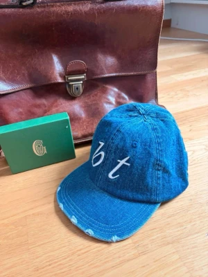 Denimkeps  Bristol textiles - Säljer en snygg blå keps i denim med böjd skärm och vit broderad text 'bt' framtill. Kepsen har klassisk sexpanelsdesign och justerbar rem bak. Perfekt för dig som gillar streetwear och vill ha en avslappnad look.