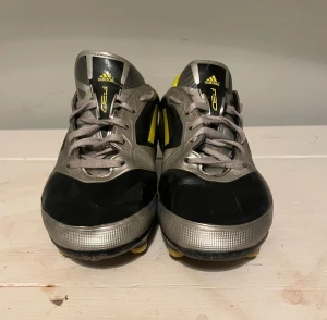 Adidas F50 fotbollsskor svart/silver - Snygga Adidas F50 fotbollsskor i svart och silver med gula detaljer och klassiska tre ränder på sidan. Skorna har snörning, låg profil och dobbar för spel på gräs. Insidan är gul och materialet är en av syntet