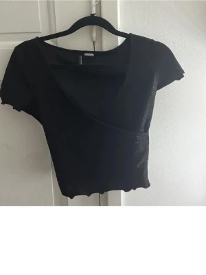 Svart topp  - Snygg svart omlottopp från Divided med korta ärmar och croppad passform. Toppen har vågig kant nertill och en djup v-ringning som ger en trendig look. Perfekt att matcha med höga jeans eller kjol.