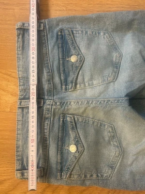 Ljusblå bootcut jeans med ficklock - Säljer ett par ljusblå bootcut jeans med snygga ficklock och silverfärgade knappar bak. Jeansen har klassisk femficksmodell och är tillverkade i denim med lätt tvättad look. Perfekta för en avslappnad och trendig stil.