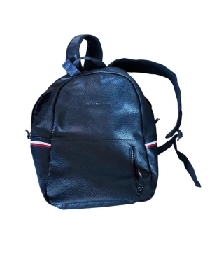Svart ryggsäck från Tommy Hilfiger - Snygg svart ryggsäck från Tommy Hilfiger i konstläder med klassiska röda, vita och blå detaljer. Ryggsäcken har justerbara axelband, dragkedjefack framtill och diskret logga på framsidan. Perfekt för skolan eller stan.