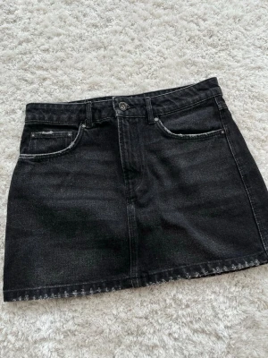 Svart jeanskjol med fickor - Säljer en svart jeanskjol i klassisk modell med fem fickor och knapp- samt dragkedjestängning framtill. Kjolen har bälteshällor och är i en kort längd, perfekt till vardagslooken. Materialet är kraftig denim som ger en snygg och tidlös känsla.