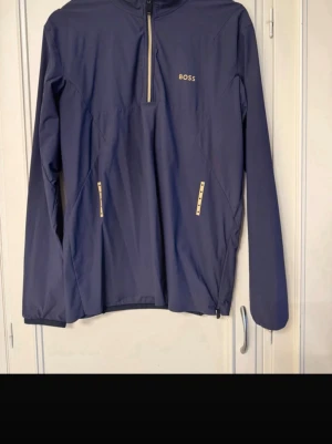 Mörkblå ziptröja från BOSS - Snygg mörkblå långärmad ziptröja från BOSS med guldfärgade dragkedjor och detaljer. Tröjan har hög krage, två fickor framtill och en halvzip som ger en sportig look. Perfekt för dig som gillar stilrena och moderna plagg.