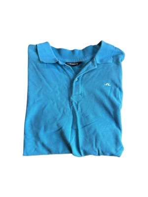 Blå golfpike från J.Lindeberg - Snygg blå pikétröja från J.Lindeberg med klassisk krage och knappar framtill. Tröjan har en liten vit logga på bröstet och är gjord i mjuk bomull som känns skön mot huden. Perfekt till sommarens alla aktiviteter.