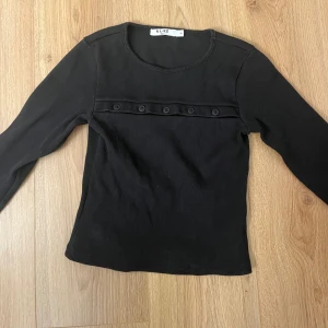 Svart ribbad topp från Chloes kollektion med  NA-KD - Svart långärmad topp från chloes kollektion med NA-KD i ribbad bomull med rund halsringning. Dekorerad med en rad svarta knappar framtill över bröstet. Enkel och clean design som funkar till många olika stilar.