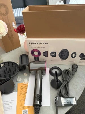 Dyson Supersonic hårfön med tillbehör - Säljer en Dyson Supersonic hårfön i metallicgrå och lila med flera stylingmunstycken. Medföljer diffuser, stylingkoncentrator och andra tillbehör för olika hårtyper. Snygg och modern design, perfekt för dig som vill ha ett proffsigt resultat hemma.