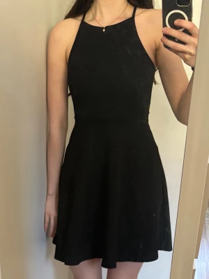 Svart ärmlös kortklänning - Säljer en svart, enkel kortklänning med hög hals och smala axelband. Klänningen har en figurnära överdel och utställd kjol. 