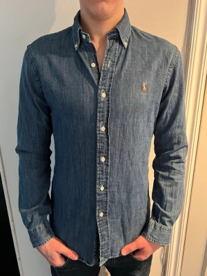 Blå jeansskjorta från Ralph Lauren - Klassisk blå jeansskjorta från Ralph Lauren med button-down krage och vita knappar. Det är inte riktigt jeans material utan lite mjukare och sitter som slimfit, modellen på bilden är 187 lång, och skjortan är i storlek M