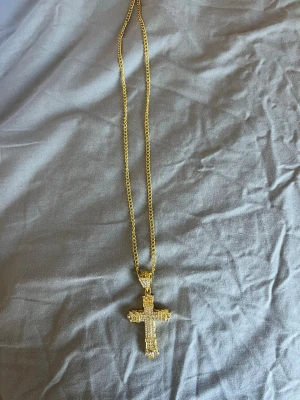 Guldfärgat halsband med korshänge - Snyggt halsband i guldfärg med ett stort korsformat hänge dekorerat med glittrande stenar. Kedjan är medelgrov och hänget har detaljerade kanter som ger ett exklusivt intryck. Perfekt accessoar för dig som vill sticka ut med något unikt och trendigt.