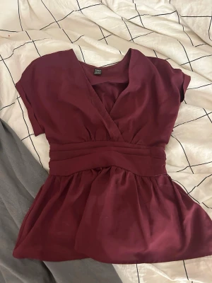 Vinröd omlottopp med markerad midja - Snygg vinröd omlottopp med v-ringning och markerad midja. Toppen har korta ärmar och ett lätt utställt fall nedtill. Perfekt att styla med jeans eller kjol för en trendig look.