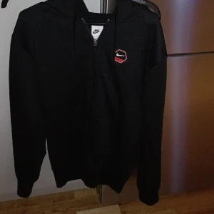 Svart Nike hoodie med röda detaljer riktigt snygg hoddie  - Svart hoodie från Nike med huva och snörning. Stora Nike-tryck i silver på ryggen och röda band med Nike-logga längs ärmarna. Dragkedja framtill och liten Nike-logga på bröstet. Perfekt för dig som gillar sportig och clean stil. Endast använda testad hemma jöpt för 1200 säljes för 600
