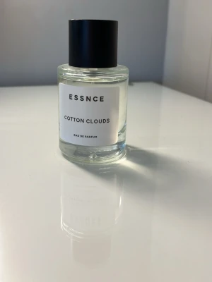 ESSNCE Cotton Clouds Eau de Parfum - Minimalistisk parfymflaska från ESSNCE med doften Cotton Clouds. Flaskan är cylindrisk i genomskinligt glas med svart rund kork och vit etikett med svart text. En stilren och modern parfym som passar perfekt i badrumshyllan.