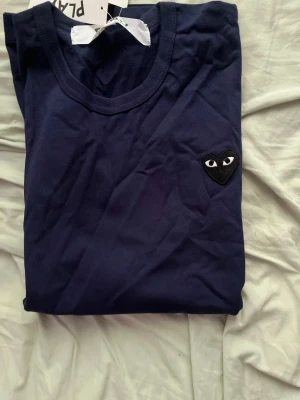 Mörkblå t-shirt Comme des Garçons Play - Säljer en mörkblå t-shirt från Comme des Garçons Play med det ikoniska hjärta-logotypen på bröstet. T-shirten är i mjuk bomull och har en klassisk rund hals och korta ärmar. Perfekt för dig som gillar stilrena plagg med en twist.
