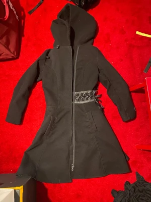 Svart kappa med snörning och huva - varm kappa med stor huva och dragkedja framtill. Figursydd modell med snörning i midjan både fram och bak. snörningen där bak gör att man verkligen kan få den snatched. passar stl s-m