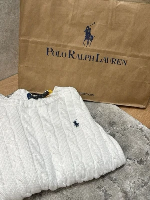 Vit kabelstickad tröja Polo Ralph Lauren - Kabelstickad vit tröja från Polo Ralph Lauren med rund halsringning och den klassiska broderade loggan i marinblått på bröstet. Tröjan har ribbade muddar vid ärmslut och nederkant. Perfekt för en clean och stilren look.