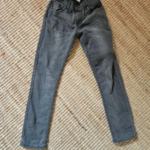 Gråa skinny jeans från Levi's stl 12A - Snygga gråa skinny jeans från Levi's med klassisk femficksdesign och dragkedjegylf. Jeansen har en smal passform och subtila slitningar framtill för en cool look. Perfekta till sneakers eller boots.