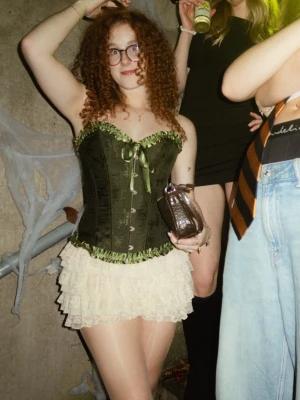 Grön korsett med spetsdetaljer - Säljer en snygg grön korsett med satinband och spetsdetaljer. Den har en figurnära passform och knäppning framtill. Har endast använt en gång