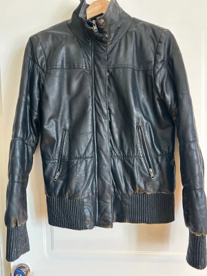 Svart skinnjacka från Object - Snygg svart skinnjacka från Object med hög krage och dragkedja framtill. Jackan har två framfickor med dragkedja och ribbade muddar vid ärmslut och nederkant för extra stil. Perfekt för dig som vill ha en klassisk men cool look.