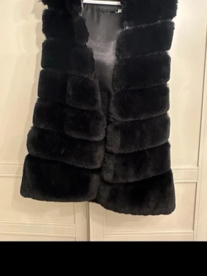 Svart fluffig pälsväst - Säljer en svart, mjuk och fluffig pälsväst med breda horisontella paneler. Västen är ärmlös och har en öppen front utan knappar eller dragkedja. Perfekt för att addera en lyxig touch till din outfit.