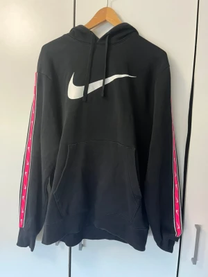 Svart Nike hoodie med rosa detaljer - Snygg svart hoodie från Nike med stor vit logga på bröstet och rosa band med små vita swoosh längs ärmarna. Klassisk känguruficka framtill och justerbar huva med snören. Perfekt för en sportig och avslappnad stil.
