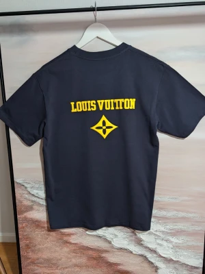 Iouis Vuitton T-shirt  - Mörkblå t-shirt i bomull med gult tryck. Framsidan pryds av en centrerad logotyp, och baksidan har ett större texttryck tillsammans med en grafisk blomstermodul. Tröjan har en klassisk passform med rund halsringning och korta ärmar.( Fast pris)
