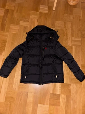 Svart pufferjacka från Polo Ralph Lauren - Snygg svart pufferjacka från Polo Ralph Lauren med huva och dragkedja framtill. Jackan har två framfickor med dragkedja och en liten röd logga på bröstet. Tillverkad i glansigt material som ger en modern look och håller dig varm under kalla dagar.