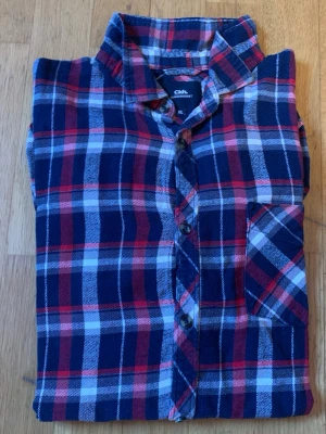 Rutig skjorta från Clockhouse XL - Snygg rutig skjorta från Clockhouse i blått, rött och vitt. Skjortan har klassisk krage, knäppning framtill och en bröstficka. Perfekt för en avslappnad stil och tillverkad i mjuk bomull.