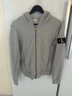 Grå hoodie från Calvin Klein Jeans - Snygg grå hoodie med dragkedja från Calvin Klein Jeans. Hoodien har en stor huva, ribbade muddar och fickor framtill. På ena ärmen sitter en svart patch med CK-logga i vitt. Tillverkad i mjukt sweatshirtmaterial, perfekt för en avslappnad look.