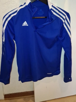 Blå Adidas träningströja Aeroready - Snygg blå långärmad träningströja från Adidas med vita ränder på axlarna och dragkedja vid halsen. Tillverkad i funktionsmaterialet Aeroready som andas bra. Perfekt för träning eller uppvärmning. Adidas-logga på bröstet.