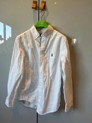 Vit skjorta från Ralph Lauren - Klassisk vit skjorta från Ralph Lauren med button-down krage och broderad logga på bröstet. Skjortan är långärmad och tillverkad i mjuk bomull, perfekt för en clean och stilren look.