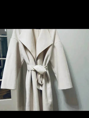 Kappa från nelly x Bianca storlek 36 - Stilren ljusbeige kappa med bred krage och knytskärp i midjan. Kappan har en rak passform och långa ärmar, perfekt för att skapa en elegant look. Tillverkad i ett mjukt tyg som ger en lyxig känsla.