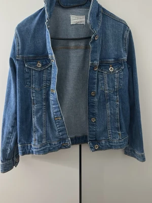 Klassisk blå jeansjacka - Snygg blå jeansjacka med raka ärmar och två bröstfickor med knapp. Jackan har silverfärgade knappar framtill och på ärmsluten. Tidlös modell som passar till det mesta och har en avslappnad passform. Perfekt att slänga över en hoodie eller t-shirt.