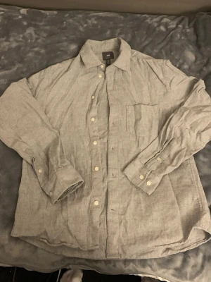 Ljusgrå linneskjorta från H&M - Säljer en stilren ljusgrå skjorta från H&M i linne. Skjortan har klassisk krage, långa ärmar med manschett och vita knappar framtill samt en bröstficka. Perfekt för dig som gillar en avslappnad men snygg look.