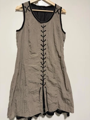 Beige ärmlös klänning Wearhouse - Säljer en beige ärmlös klänning från Wearhouse med svarta dekorativa snörningsdetaljer framtill och runt ärmhålen. Klänningen har en avslappnad passform och svarta kontrastkanter. Perfekt för dig som gillar unika detaljer och enkel stil.