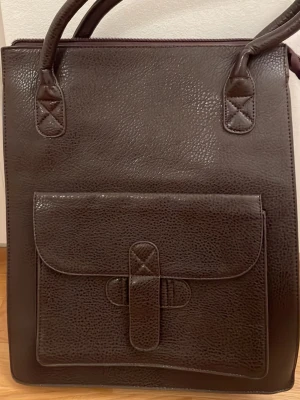 Mörkbrun fyrkantig handväska - Snygg mörkbrun handväska i konstläder med fyrkantig form och dubbla handtag. Väskan har en stor ficka framtill med lock och spänne samt en mindre ficka med dragkedja på baksidan. Perfekt för dig som gillar stilrena och praktiska accessoarer.