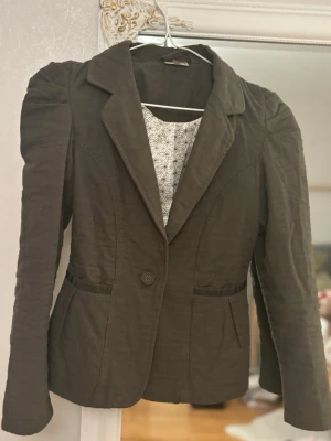 Mörkbrun kavaj med puffärm - Snygg mörkbrun/grön  kavaj med puffade axlar och figurnära passform från POMP DE LUX. Kavajen har ett klassiskt slag, en knapp framtill och två fickor. Fodret har ett grafiskt mönster i vitt och svart. Perfekt för dig som vill ha en unik twist på din outfit.