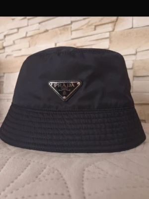 Svart bucket hat från Prada - Snygg svart bucket hat från Prada med den ikoniska metalltriangeln framtill. Hatten är tillverkad i ett glansigt nylonmaterial som ger en modern och clean look. Perfekt accessoar för att lyfta din outfit med en exklusiv touch.
